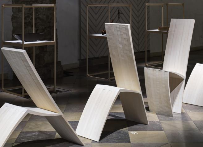 Muzeja telpā izstādīti trīs balti, skulpturāli krēsli ar izliektām, ģeometriskām formām, fonā redzamas vitrīnas ar dizaina priekšmetiem.