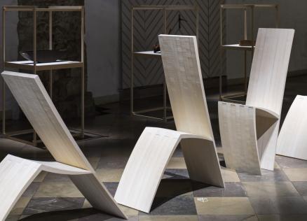 Muzeja telpā izstādīti trīs balti, skulpturāli krēsli ar izliektām, ģeometriskām formām, fonā redzamas vitrīnas ar dizaina priekšmetiem.