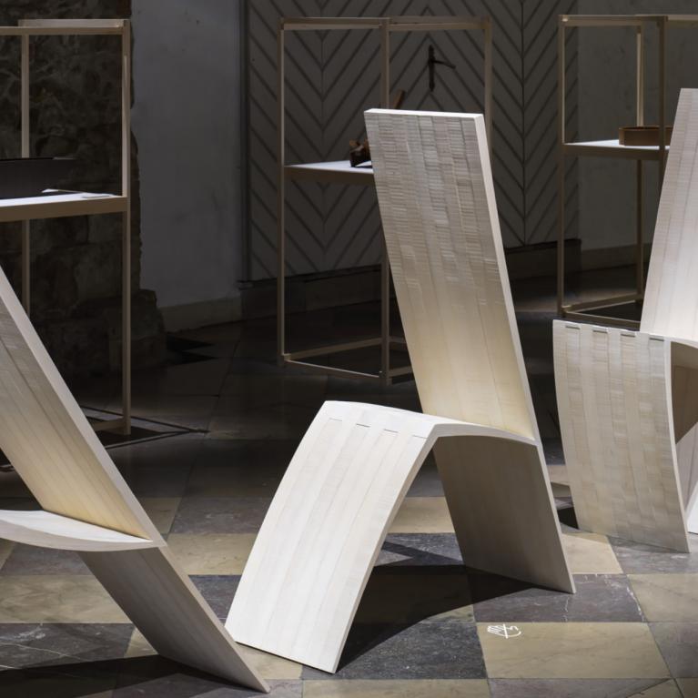 Muzeja telpā izstādīti trīs balti, skulpturāli krēsli ar izliektām, ģeometriskām formām, fonā redzamas vitrīnas ar dizaina priekšmetiem.