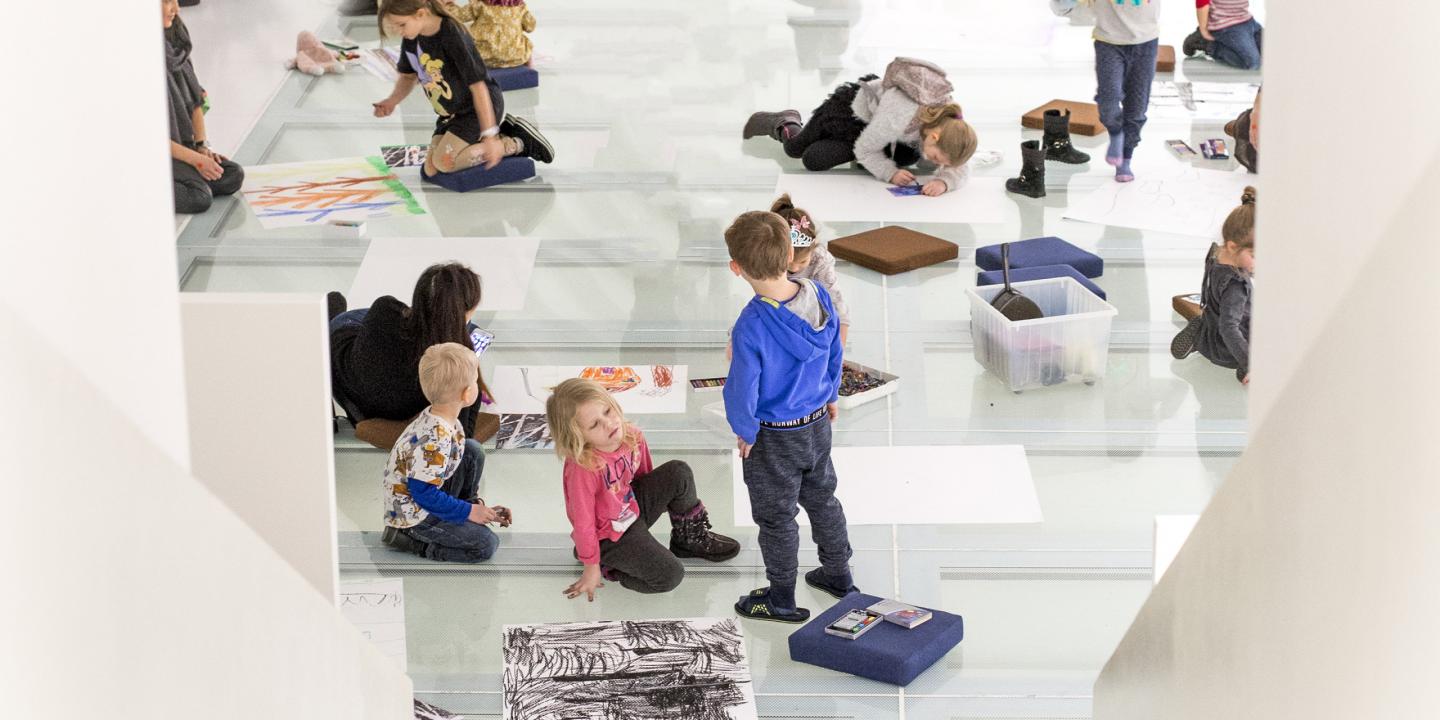 Bērni sēž un zīmē uz stiklotas grīdas muzeja radošajā nodarbībā, apkārt izklātas lapas un krītiņi.