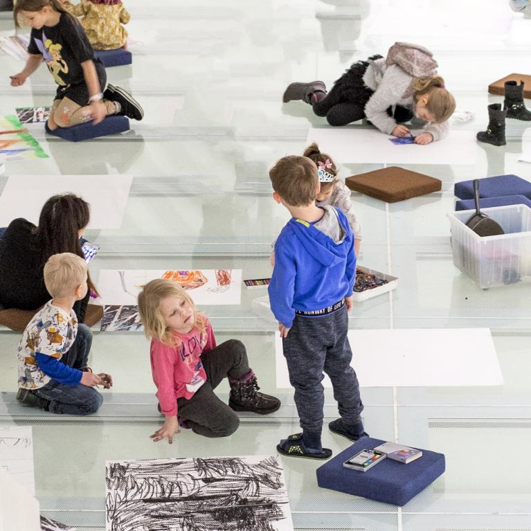 Bērni sēž un zīmē uz stiklotas grīdas muzeja radošajā nodarbībā, apkārt izklātas lapas un krītiņi.