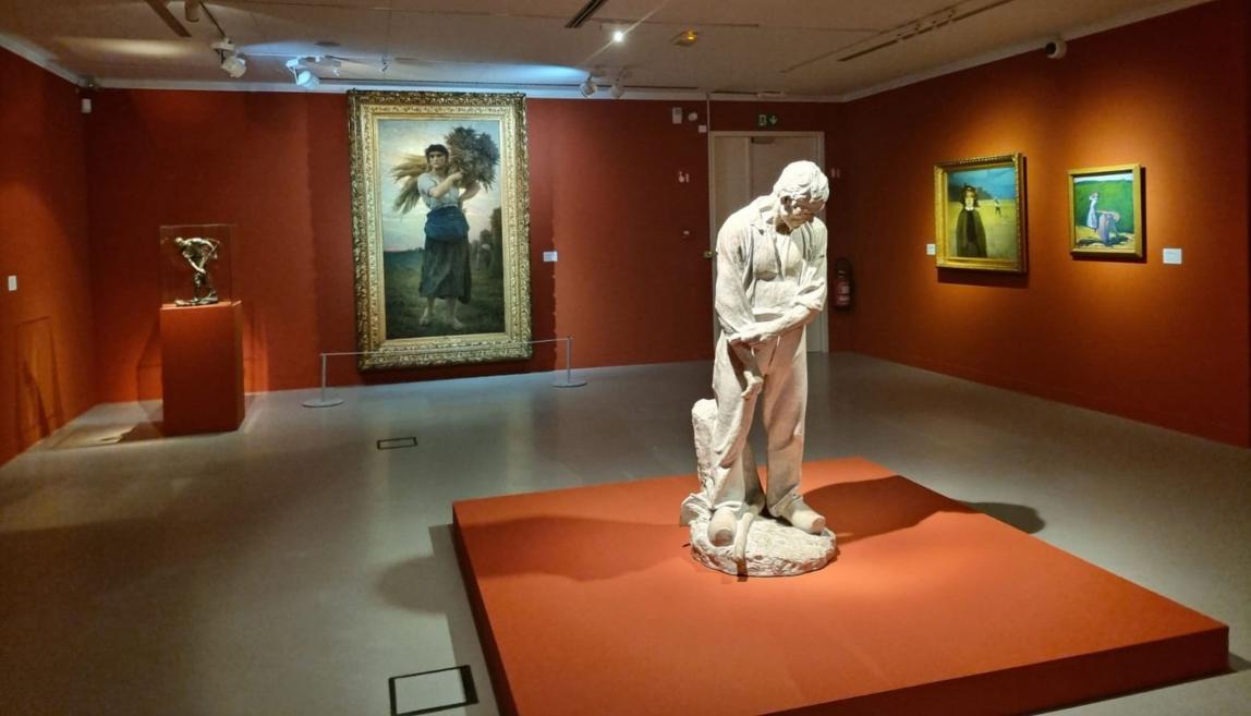 Izstādes zāle ar gleznām un skulptūrām uz sarkanām sienām, priekšplānā balta lauku strādnieka skulptūra.