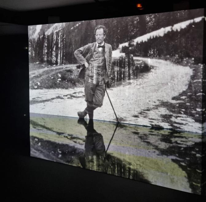 Melnbalts fotoprojekcijas kadrs ar vīrieti, kas atbalstās uz spieķa lauku ceļā.