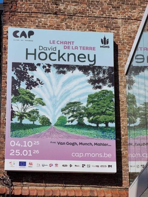 Ēkas fasāde ar izstādes plakātu “David Hockney. Le Chant de la Terre” uz ķieģeļu sienas.