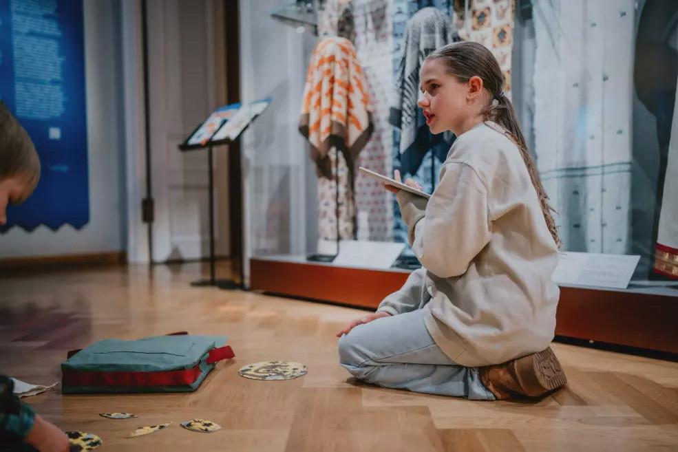 Meitene sēž uz grīdas pie muzeja ekspozīcijas un pieraksta novērojumus, blakus atrodas soma ar spēles elementiem.