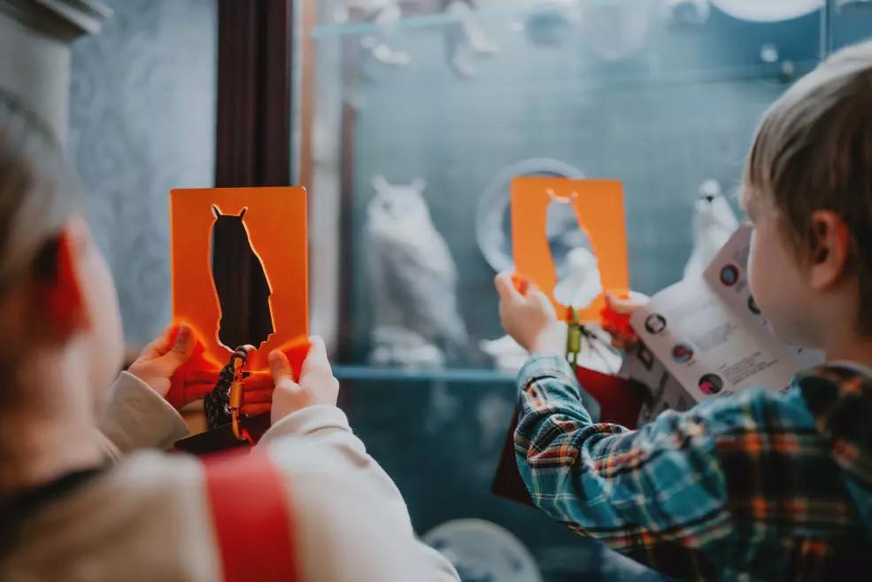 Divi bērni ar oranžām kartītēm rokās meklē ekspozīcijā priekšmetus, kas atbilst siluetiem.