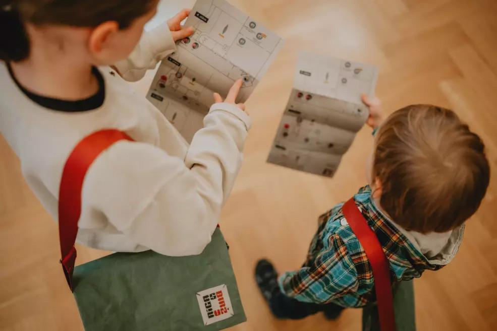 Bērni ar muzeja kartēm rokās meklē nākamo ekspozīcijas apmeklējuma punktu.
