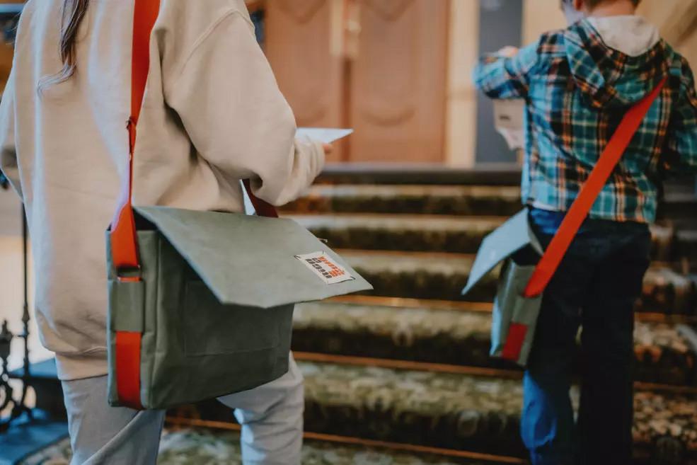 Divi bērni ar somām uz pleca kāpj pa muzeja kāpnēm, rokās turot uzdevumu lapas.