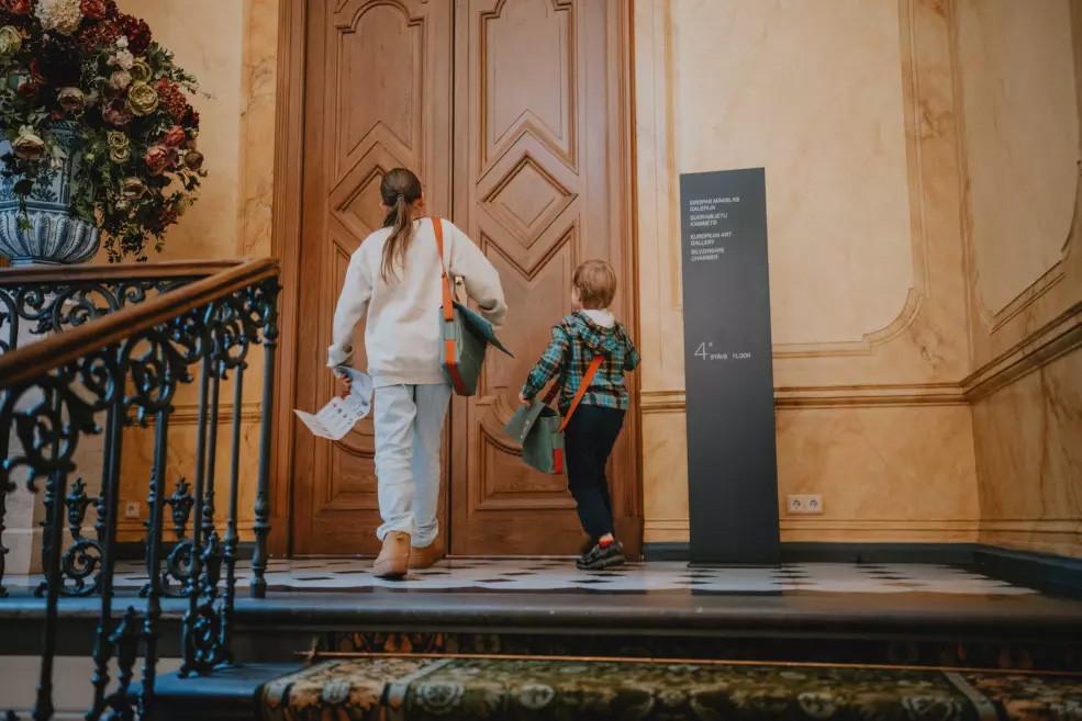 Bērni dodas augšup pa muzeja kāpnēm pret lielām koka durvīm, virs kurām redzams norāžu stabs ar ekspozīciju nosaukumiem.