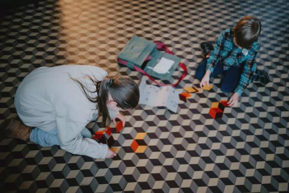 Divi bērni sēž uz ģeometriska raksta grīdas un saliek krāsainus figūru elementus, darbojoties ar muzeja somas saturu.
