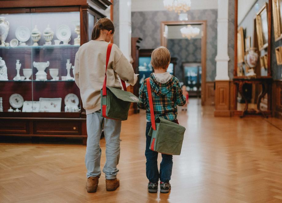 Divi bērni ar zaļām somām uz pleca apskata porcelāna un mākslas darbu ekspozīciju Latvijas Nacionālā mākslas muzeja interjerā.