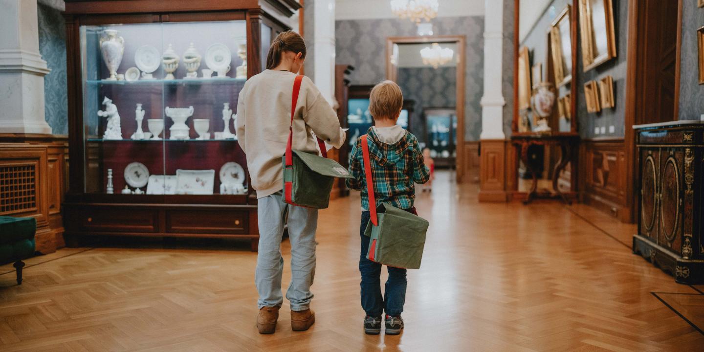 Divi bērni ar zaļām somām uz pleca apskata porcelāna un mākslas darbu ekspozīciju Latvijas Nacionālā mākslas muzeja interjerā.