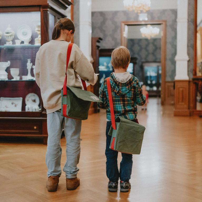 Divi bērni ar zaļām somām uz pleca apskata porcelāna un mākslas darbu ekspozīciju Latvijas Nacionālā mākslas muzeja interjerā.