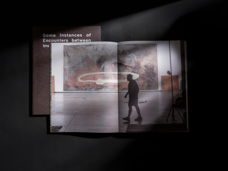 Uz melna fona novietota atvērta grāmata, kurā redzams liels darbnīcas interjera attēls ar cilvēka siluetu.