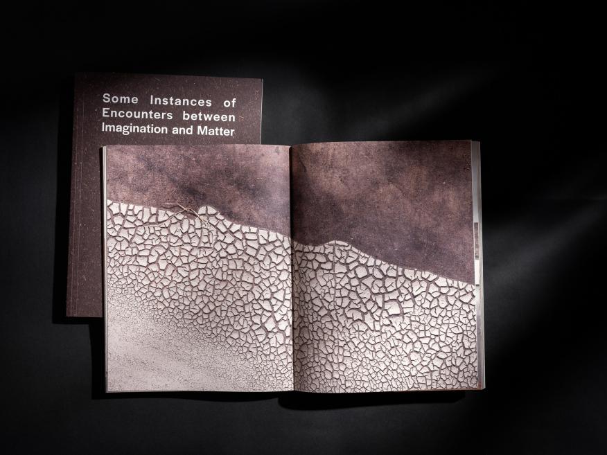 Uz melna fona novietota atvērta grāmata ar divu lappušu fotogrāfiju, kurā attēlotas zemes plaisas un faktūras.