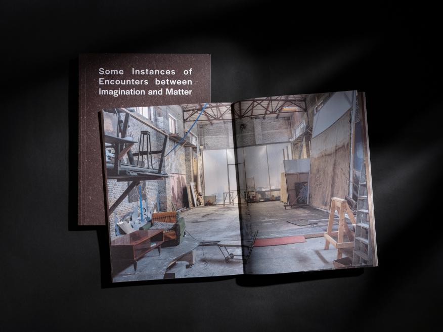 Uz melna fona novietota atvērta grāmata ar plašas, iekārtotas darbnīcas interjera fotogrāfiju.