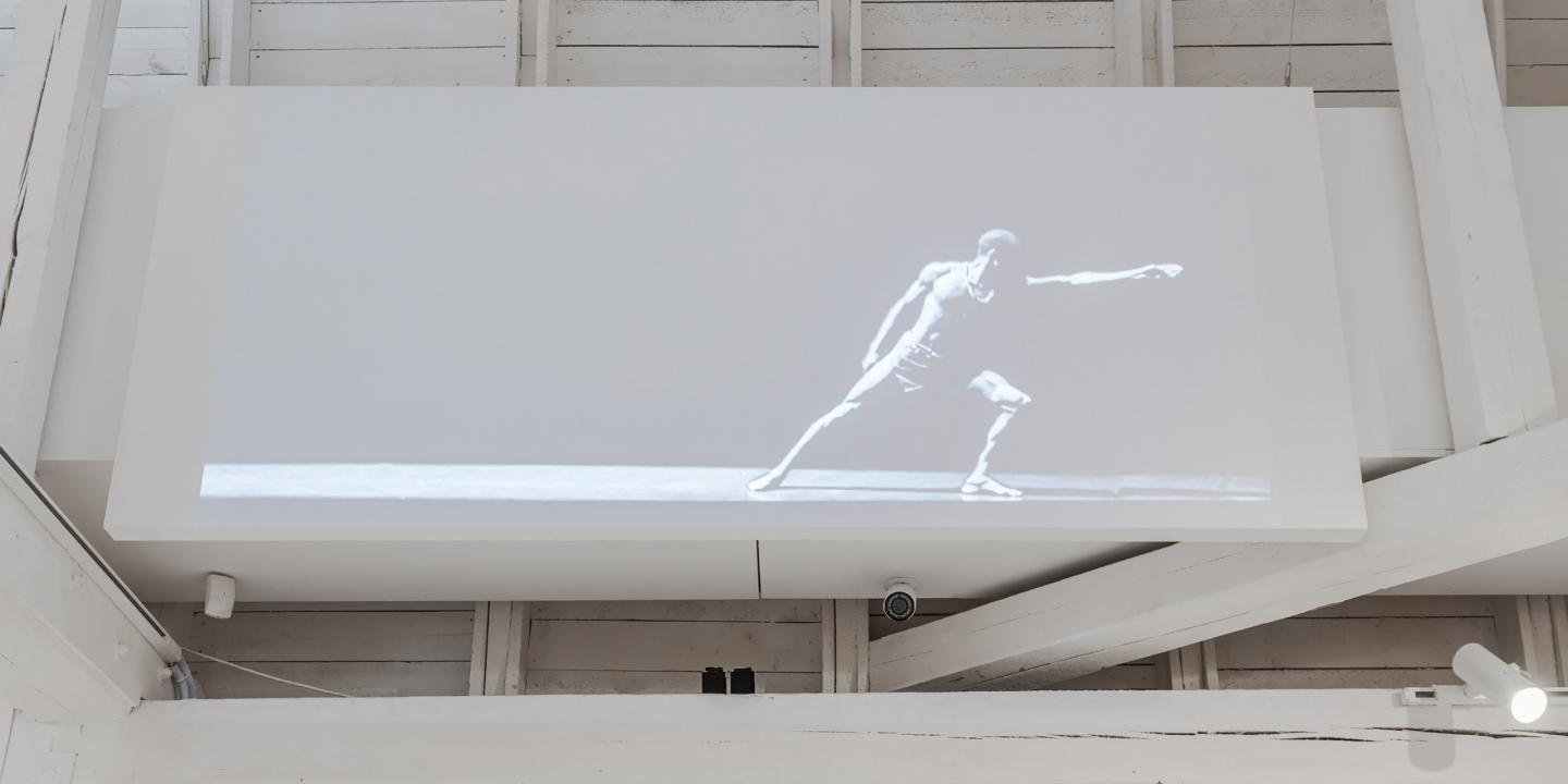 Melnbalts video projekcijas kadrs ar dejotāju dinamiskā pozā, izstādīts uz balta ekrāna modernā galerijas telpā ar baltām koka sijām.