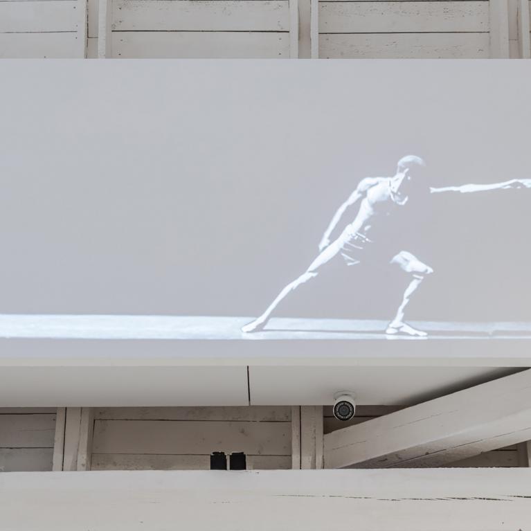 Melnbalts video projekcijas kadrs ar dejotāju dinamiskā pozā, izstādīts uz balta ekrāna modernā galerijas telpā ar baltām koka sijām.