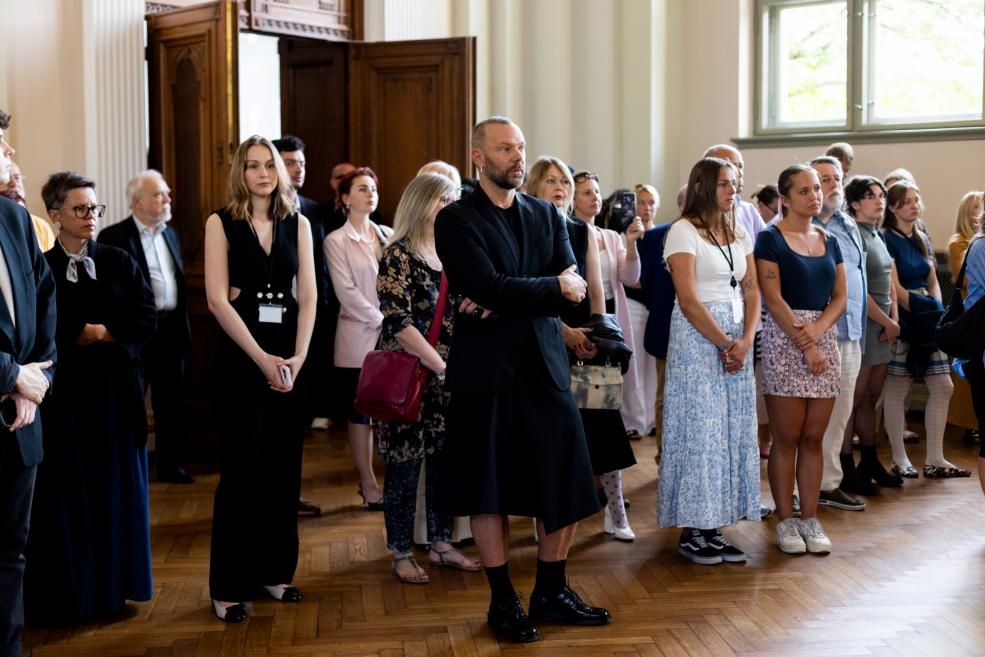 Stāvā klausītāju grupa ar nopietniem skatieniem piedalās svinīgā pasākumā vēsturiskā zālē.