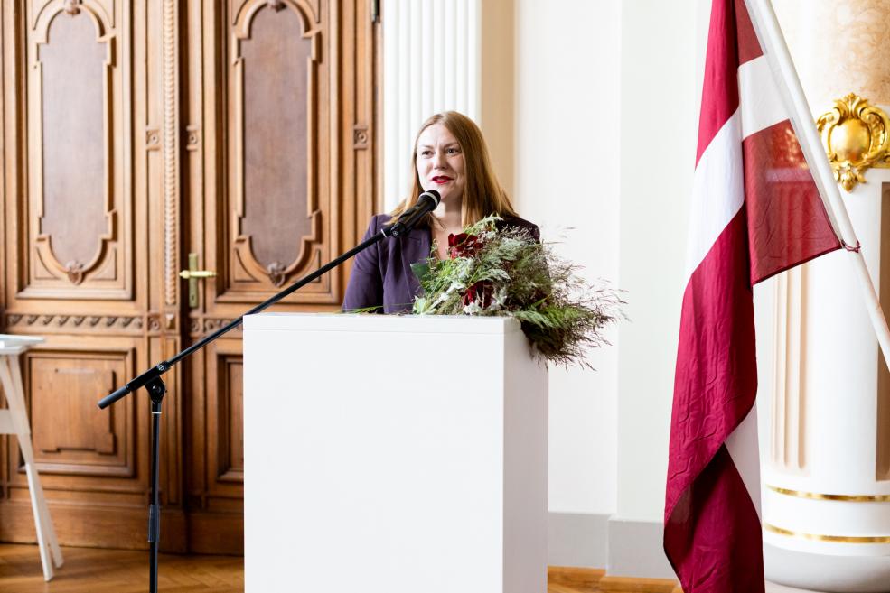 Sieviete runā no tribīnes ar mikrofonu, blakus redzams Latvijas karogs un ziedu kompozīcija.