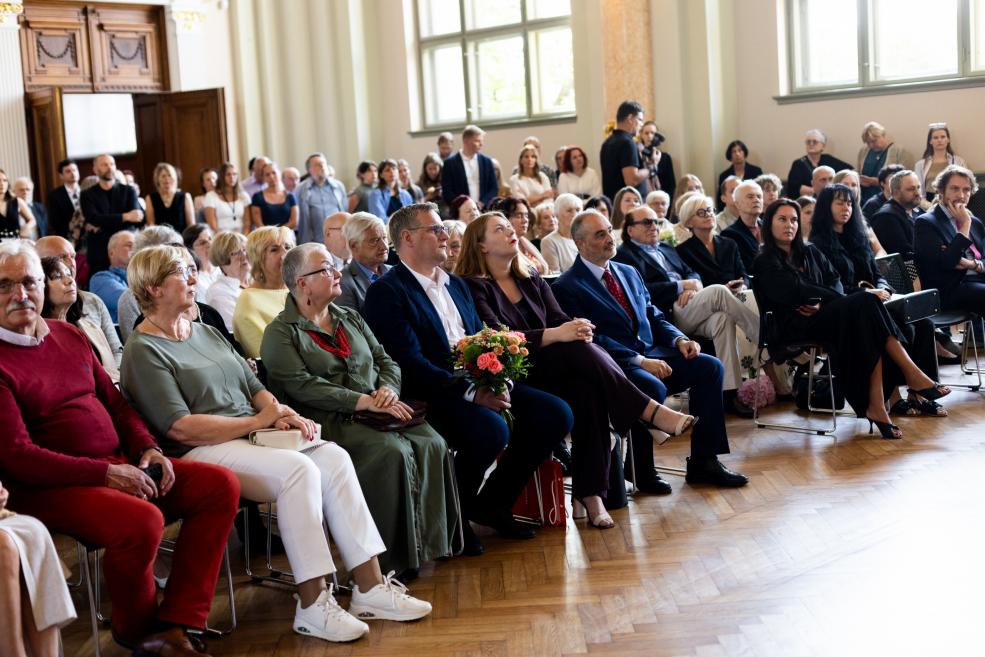 Apmeklētāji sēž svinīgā pasākumā plašā zālē, priekšplānā redzama sieviete ar ziedu pušķi.