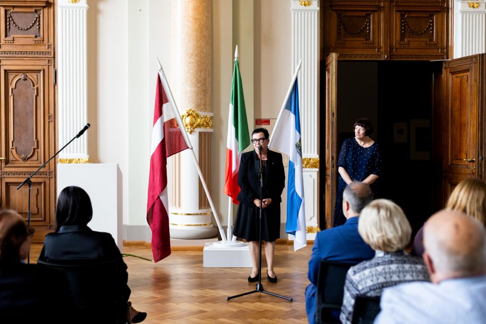 Sieviete runā mikrofonā skatītāju priekšā, fonā redzami Latvijas, Itālijas un Somijas karogi.