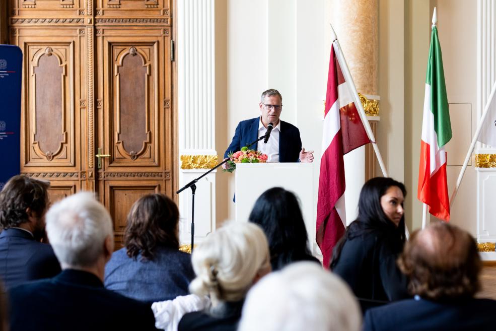 Vīrietis pie tribīnes uzstājas auditorijas priekšā, fonā redzami Latvijas un Itālijas karogi.