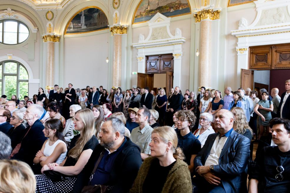 Liela cilvēku auditorija sēž un stāv greznā zālē, uzmanīgi sekojot pasākuma norisei.