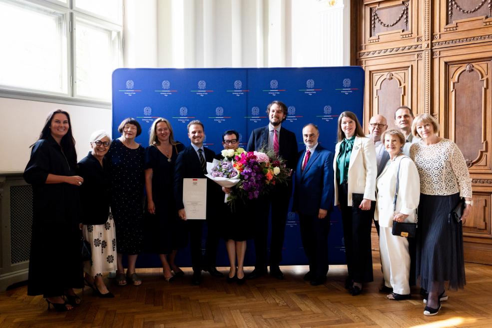 Grupas foto pie oficiāla fona, centrā sieviete ar ziedu pušķi un apliecinājuma dokumentu rokās.
