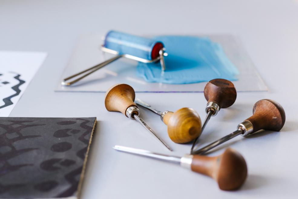 Uz galda novietoti linogriezuma instrumenti ar koka rokturiem, priekšplānā iespiedforma, fonā krāsas rullītis ar zilu krāsu.