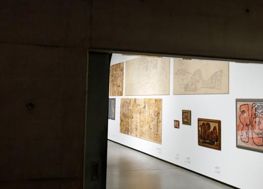 Skats no telpas uz muzeja galeriju, kur baltā sienā izstādīti dažādi zīmējumi un gleznas dažādos izmēros.