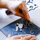 Tuvplānā redzamas rokas, kas ar instrumentu veido zīmējumu, skrāpējot līnijas tumšā virsmā.