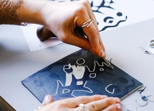 Tuvplānā redzamas rokas, kas ar instrumentu veido zīmējumu, skrāpējot līnijas tumšā virsmā.