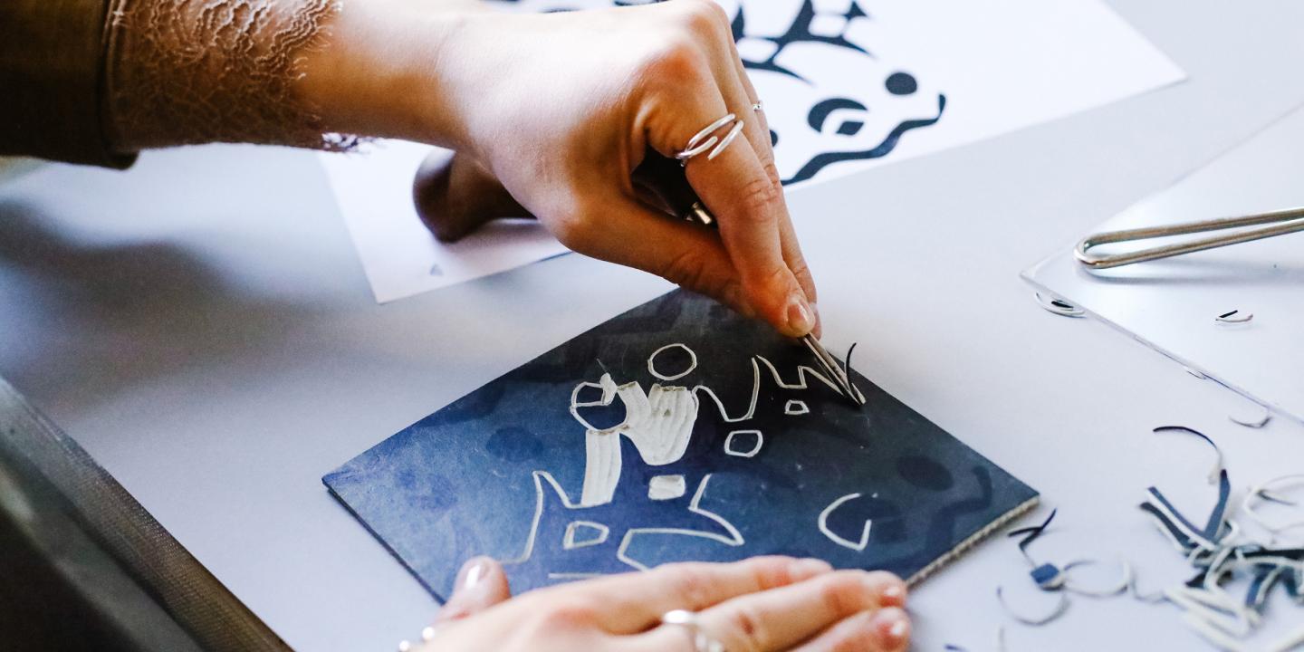 Tuvplānā redzamas rokas, kas ar instrumentu veido zīmējumu, skrāpējot līnijas tumšā virsmā.