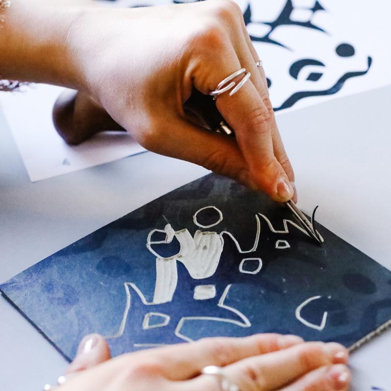 Tuvplānā redzamas rokas, kas ar instrumentu veido zīmējumu, skrāpējot līnijas tumšā virsmā.