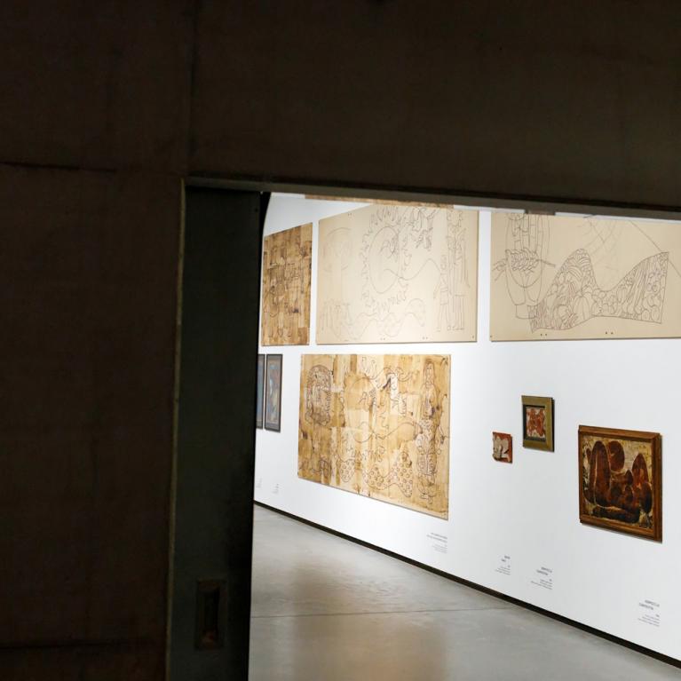 Skats no telpas uz muzeja galeriju, kur baltā sienā izstādīti dažādi zīmējumi un gleznas dažādos izmēros.