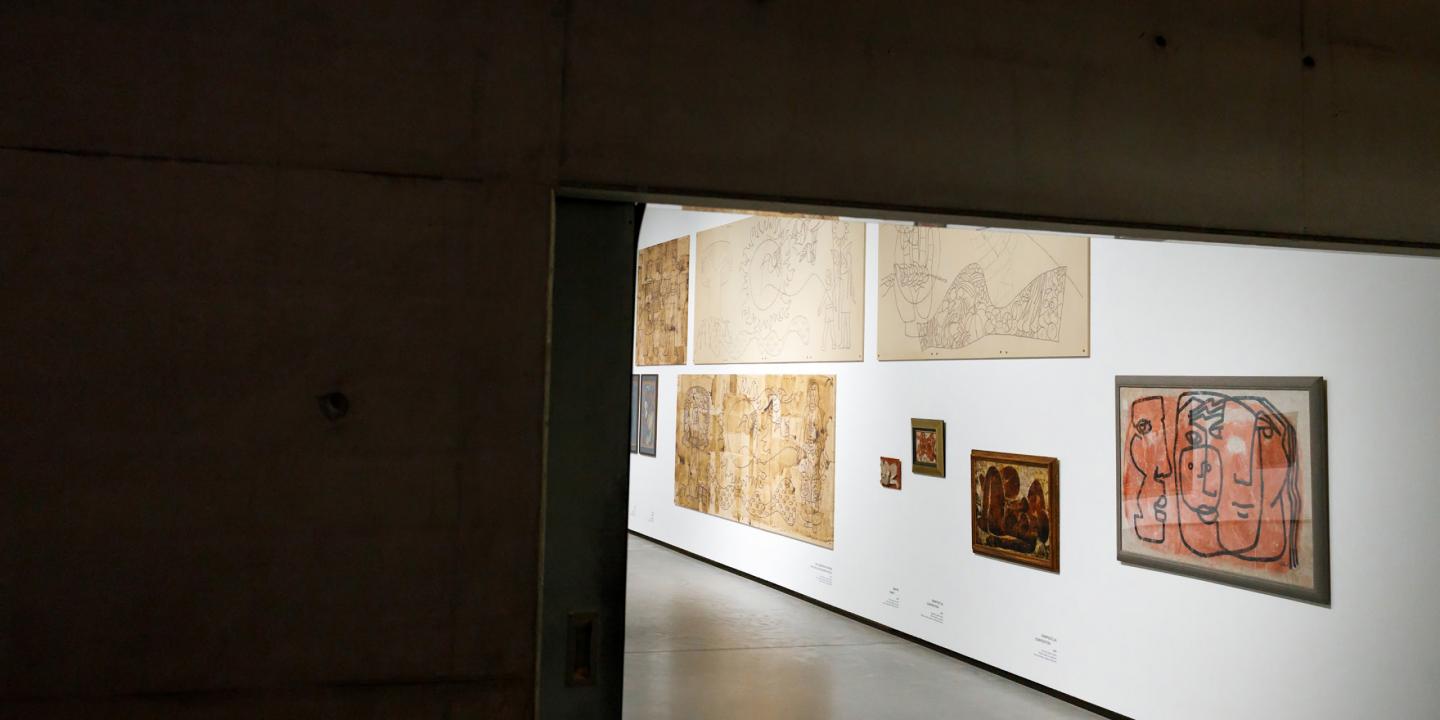 Skats no telpas uz muzeja galeriju, kur baltā sienā izstādīti dažādi zīmējumi un gleznas dažādos izmēros.